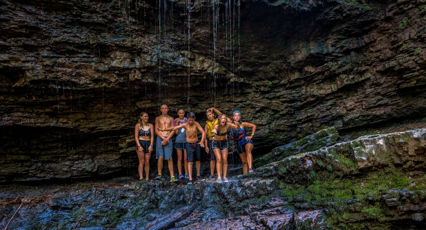 A group of young people wearing swimwear stand on a rocky ledge under a slowly trickling waterfall. 